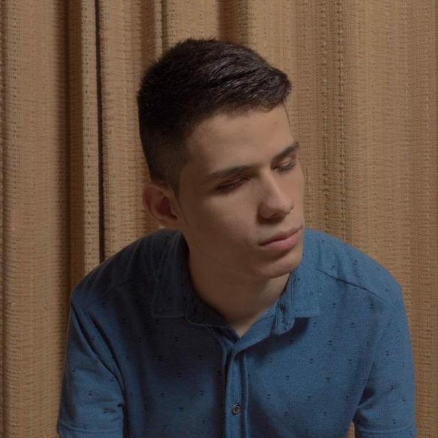 El joven tiene el cabello corto, oscuro y está peinado hacia un lado. Su rostro es de forma ovalada y su piel es clara. Sus cejas son gruesas y oscuras, y tiene ojos medianos que están entrecerrados, mirando hacia abajo o hacia la derecha, lo que da una impresión de que está reflexionando o en un momento introspectivo. Sus labios son medianos y su boca está cerrada, con una ligera expresión seria.  Está vistiendo una camisa tipo polo de color azul, que tiene botones al frente y pequeños puntos o