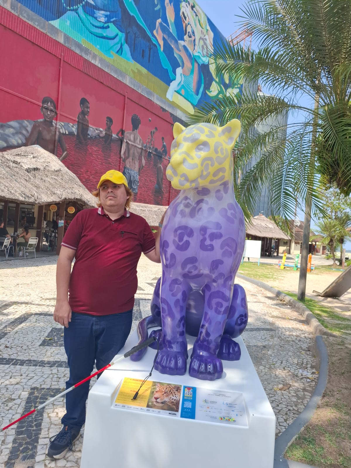 Soy yo con gorra amarilla, camiseta tipo polo color burdeos, geans azul, zapatillas azules con blanco, sostengo bastón en una mano y apoyo mi mano sobre la estatua de un jaguar. De fondo hay un mural y palmeras.