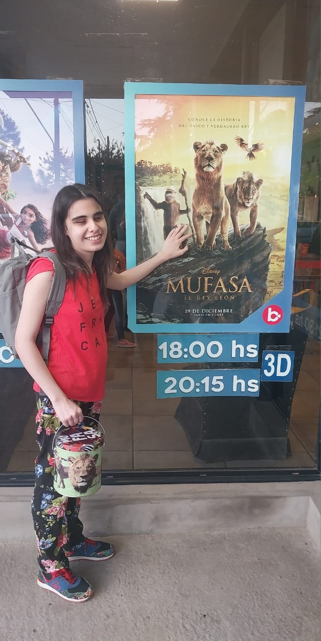 soy yo, Sizi, en la puerta de un cine. Mi cabello es largo, castaño y está suelto, volando por ahí. Tengo puesta  una remera roja, pantalón estampado con flores y unas zapatillas de colores. En mi espalda llevo mi mochila, que es Gris. Y en mi mano derecha sostengo con orgullo un balde de palomitas decorado con la cara de Mufasa. Estoy sonriendo feliz, y con mi mano izquierda estoy tocando con respeto el cartel de la película "Mufasa: El Rey León".