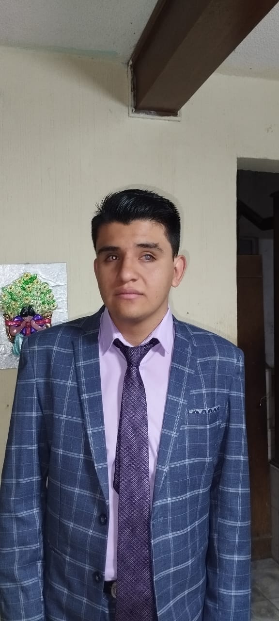 Soy yo, de piel clara y cabello oscuro peinado. Miro al frente con una expresión tranquila. Llevo una camisa lavanda, un saco azul a cuadros y una corbata morada.  Estoy de pie frente a una pared beige con una decoración floral a mi izquierda y el marco de una puerta oscura a mi derecha.