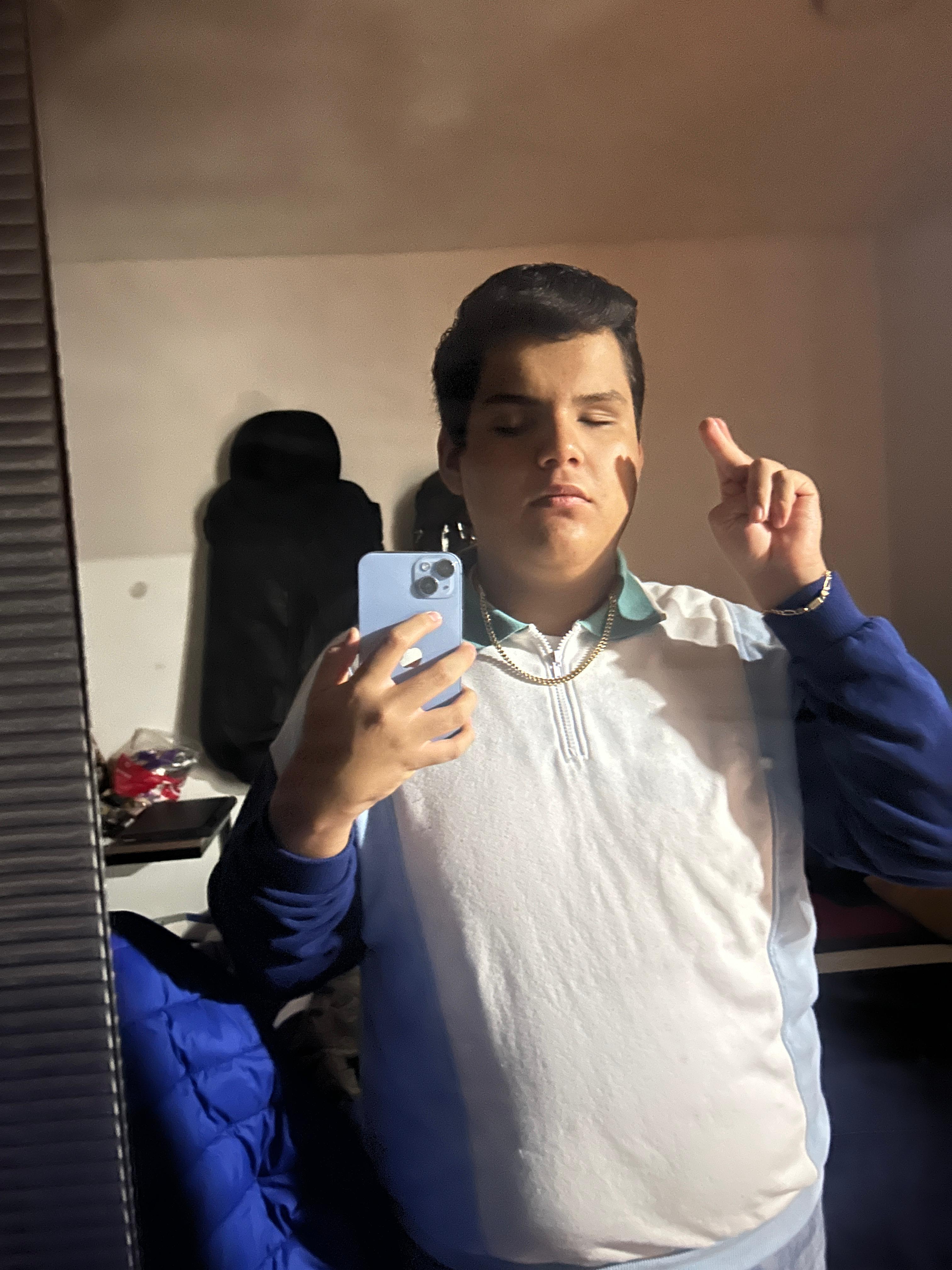 yo mirándome al espejo con una sonrisa tranquila. Llevo una sudadera polo azul claro, una cadena de oro y una esclava con mis iniciales.